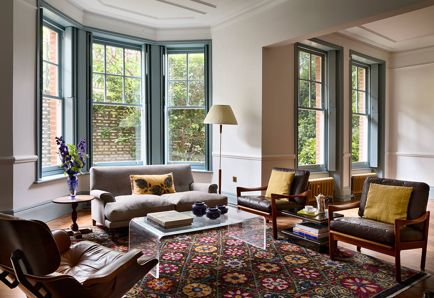 The living room of an Edwardian flat. In the centre is a patterned rug topped with a perspex coffee table, two vintage armchairs and, to the right, a grey sofa. In the background is a decorative fireplace and on its mantel rests a large gold arched mirror